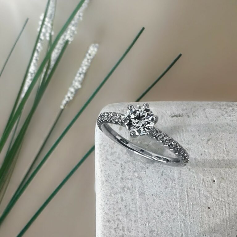 Weißgoldener Verlobungsring mit rundem Brillantsolitär und pavébesetzter Ringschiene, auf einem hellen Steinblock arrangiert vor unscharfem, beigem Hintergrund mit schlanken Gräsern.